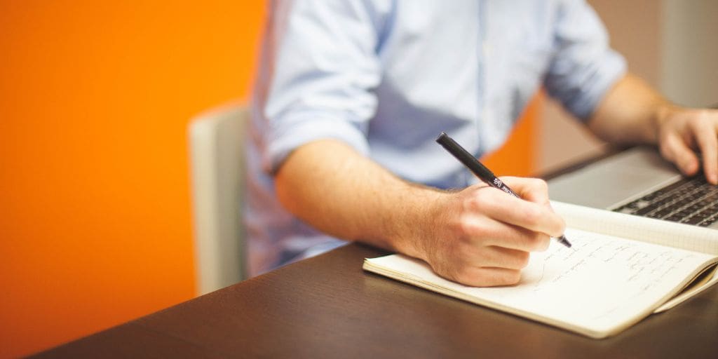 A person wearing a blue shirt writes in a notebook with a pen while using a laptop on a desk with an orange wall in the background.