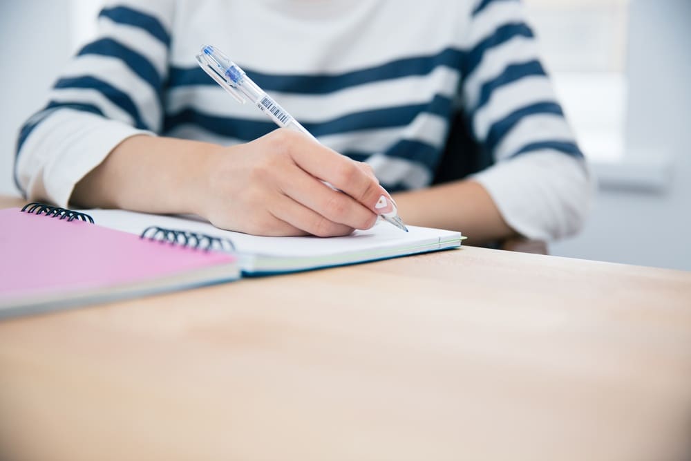 A person in a striped shirt writes in a spiral notebook with a pen at a wooden desk.