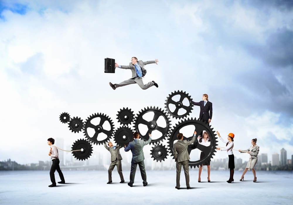 A group of people in business attire, including one in an orange helmet, are interacting with large interlocking gears. A person with a briefcase is jumping over the gears in the background.