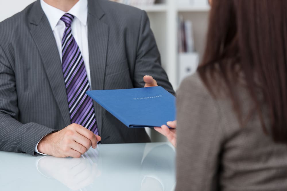 Two people in business attire are seated at a desk. One, holding a blue folder marked "Resume," is handing it to the other. Their faces are not visible in the image.