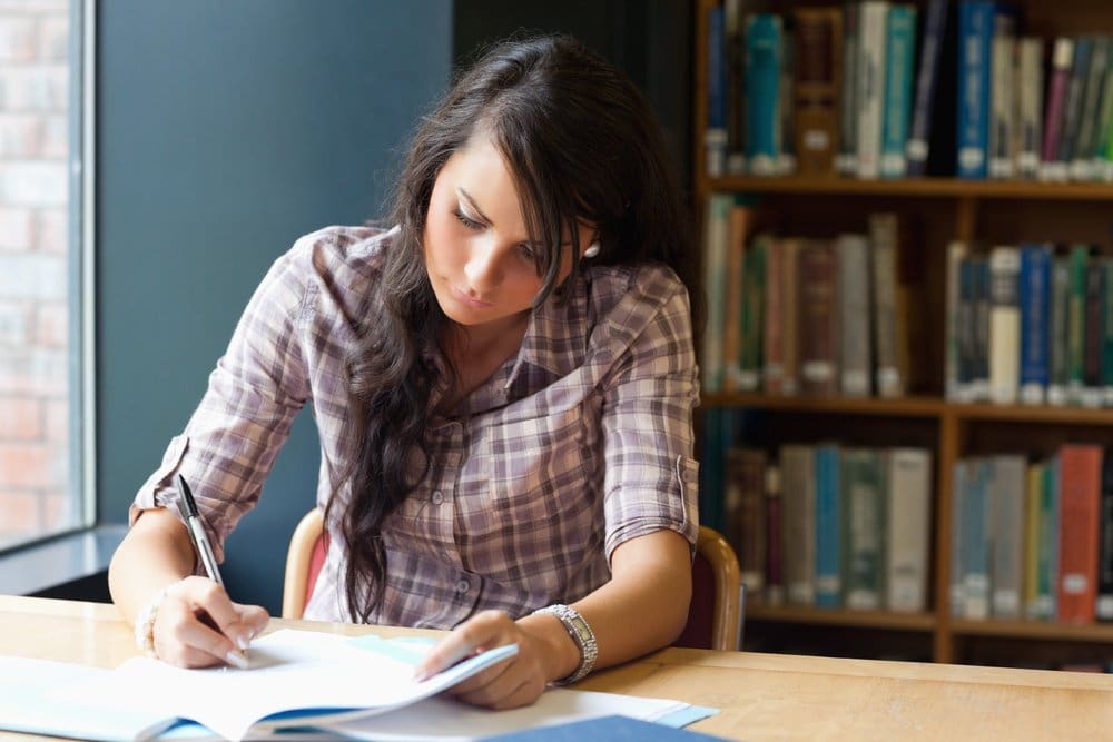 A woman with long dark hair, wearing a plaid shirt, sits at a wooden table in a library, focused on writing in a notebook.