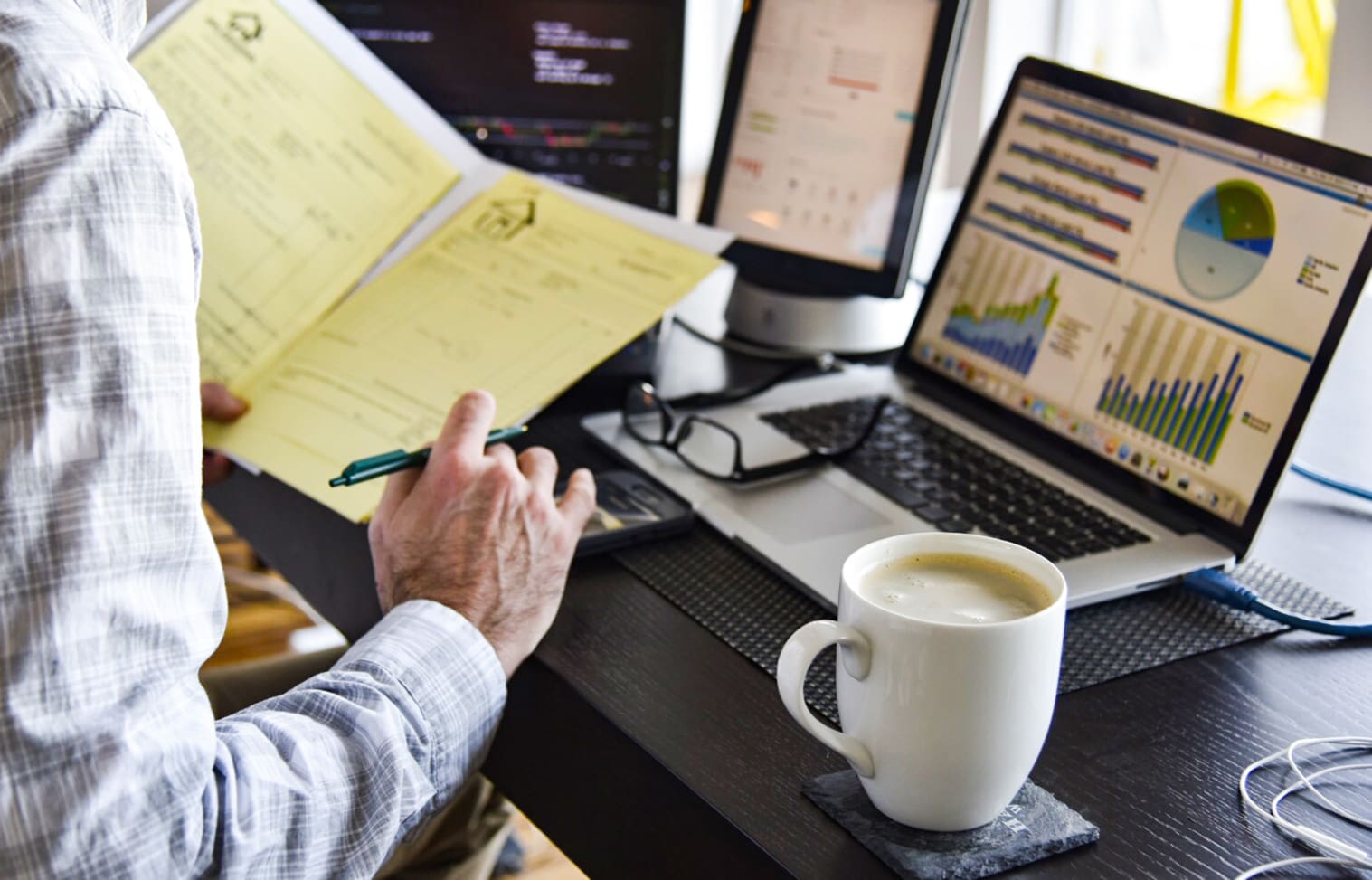 Person working on a desk with financial documents, a laptop displaying charts, an additional monitor, a calculator, and a cup of coffee.